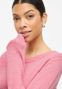 Pink sweater with ribbed neckline and cuffs, soft texture, fitted design, showcasing the model's hand near her face. Silver hoop earrings.