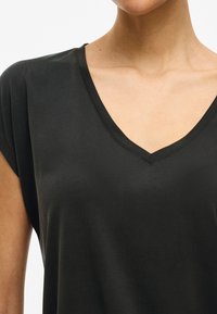 Črna T-majica z V-izrezom, narejena iz mehkega, gladkega blaga. Ima kratke rokave in sproščen kroj. Brez vzorcev ali vidnih okrasnih detajlov.