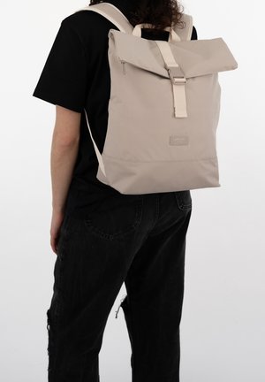 Sac à dos beige à rabat roulé avec bretelles réglables, fermeture éclair et texture lisse, porté par une personne en chemise noire.