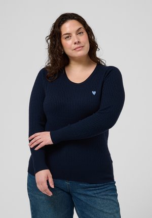 Femme aux cheveux bouclés mi-longs portant un pull à manches longues bleu marine avec un petit cœur bleu clair sur la poitrine et un jean bleu.