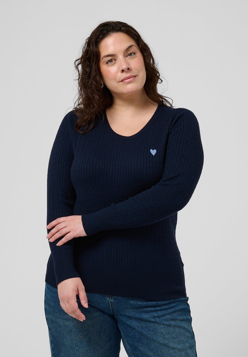 Femme aux cheveux bouclés mi-longs portant un pull à manches longues bleu marine avec un petit cœur bleu clair sur la poitrine et un jean bleu.