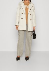 Trench-coat beige à double boutonnage avec de gros boutons, sweat-shirt marron avec détail brodé, pantalon cargo gris, et chaussures noires à bouts pointus.