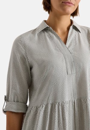 Femme portant une chemise rayée à manches longues avec col, manches retroussées, taille froncée et rayures verticales grises et blanches.