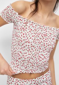 Off-the-shoulder cropped top with white base, red floral print, and green accents; smocked texture with ruffled edges.