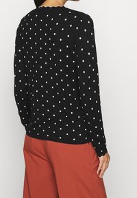 Maglione nero con pois bianchi, maniche lunghe, scollatura rotonda e bordo smerlato al collo. Texture morbida, silhouette aderente.