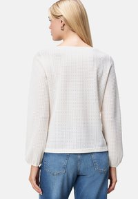 Weiße, strukturierte Bluse mit langen Ärmeln, die ein welliges Muster und einen abgerundeten Saum aufweist, kombiniert mit blauen Jeans.