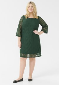 Grön spetsklänning med trekvartsärmar, prydd med blommönster och en scallopad fåll. Matcher med svarta platta skor.