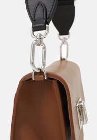 Brązowa skórzana torba o wyraźnej, sztywnej formie. Posiada srebrną sprzączkę i elementy metalowe, dopełnione czarnym, regulowanym paskiem.
