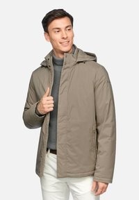 Giacca isolante marrone chiaro con cappuccio, chiusura zip frontale e tasche laterali. Indossata sopra un maglione grigio, abbinata a pantaloni chiari.