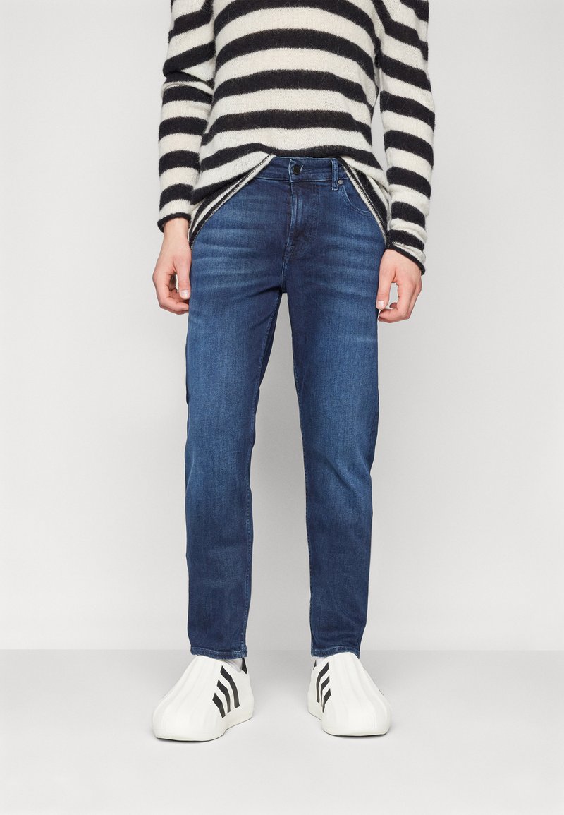 Sinised denim püksid sirge lõikega, kontrastsed õmblused ja peen kulumine. Kombineeritud musta ja valge triibulise sviitriga.