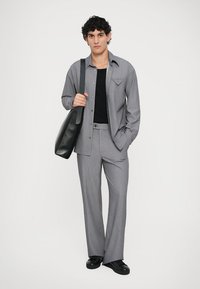 Grijze button-up overhemd met een puntzak, zwart T-shirt, grijze broek en een zwarte handtas, staand voor een witte achtergrond.