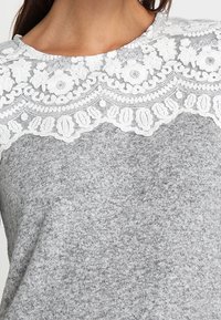 Gros plan sur un pull gris en maille avec un empiècement en dentelle blanche à motif floral et un bord festonné sur la poitrine et les épaules.