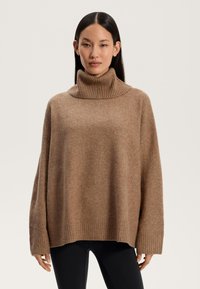Pullover marrone oversize con un colletto a imbottitura e polsini a costine, che presenta una texture morbida e una vestibilità rilassata. Pantaloni neri visibili in basso.
