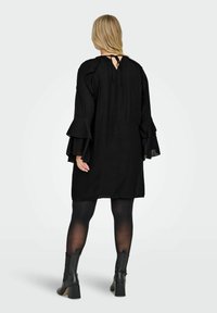 Crna haljina od lagane tkanine, s volanastim rukavima, zatvaranjem na leđima u obliku ključa i opuštenim krojem. Uparena s crnim čizmama s visokom petom.