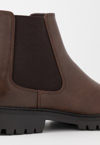 Botín Chelsea de cuero marrón con paneles laterales elásticos, suela robusta y una textura suave. Fuerte contraste entre los materiales.