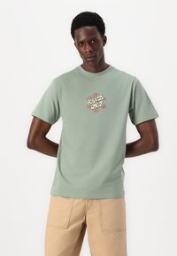 T-shirt de algodão verde claro com um gráfico circular apresentando o texto "Santa Cruz" em letras estilizadas no peito. Mangas curtas.