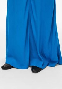 Dolga, tekoča obleka v svetlo modrem blagu; ima gladko teksturo s skritimi črnimi čevlji ob robu.