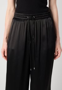 Ingrandimento di pantaloni neri in satin con vita elastica e cordino, indossati da una persona su uno sfondo neutro.