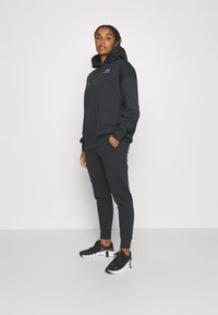 Felpa nera con cappuccio e lacci, dotata di tasca frontale e logo. Abbinata a pantaloni jogger aderenti coordinati e scarpe da ginnastica nere.