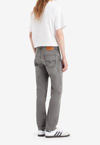Jeans in denim grigio con una vestibilità a gamba dritta, dotati di una toppa in pelle sulla cintura posteriore e cuciture a contrasto. Abbinati a una maglietta bianca e scarpe sportive.