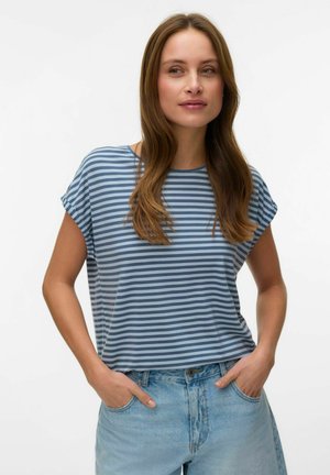 Ung kvinde med langt hår iført en blåstribet T-shirt og lyseblå jeans, stående med hænderne i lommerne mod en ensfarvet baggrund.