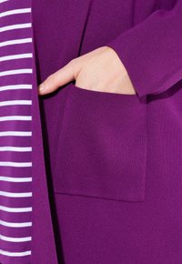 Lila gestrickte Strickjacke mit einer Vorderasche, getragen über einem gestreiften weißen und lila Hemd. Der Stoff hat eine glatte Textur und gerippte Details.