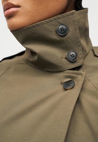 Κοντινό πλάνο ενός ατόμου που φοράει παλτό ελιάς με υψηλό γιακά, κλειστό με τρία μεγάλα σκουρόχρωμα κουμπιά.