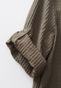 Manche de chemise texturée marron retroussée et maintenue par une lanière en tissu boutonnée sur fond blanc.