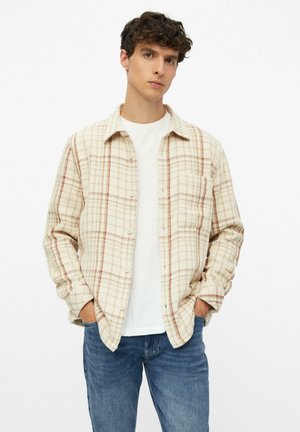 Junger Mann mit lockigem Haar, der ein cremefarbenes und braunes kariertes Hemd über einem weißen T-Shirt und blauen Jeans trägt, steht mit den Händen in den Taschen.