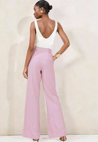 Top bianco senza maniche con scollatura a V profonda sul retro, abbinato a pantaloni rosa a vita alta e gamba larga. Il tessuto liscio e il design su misura esaltano la vestibilità.