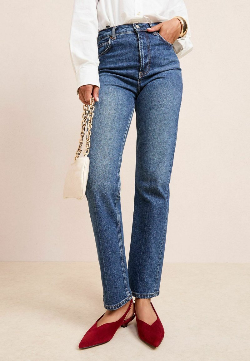 Jeans din denim albastru cu talie înaltă, cu un croi drept, asortați cu pantofi roșii din suede, tip slip-on, și o geantă mică albă texturată cu un umăr din lanț.