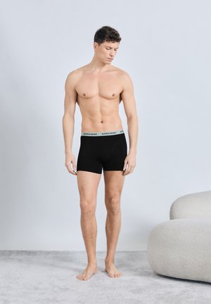 Homme torse nu portant un boxer noir Björn Borg, debout pieds nus sur un tapis clair à côté d'un canapé blanc incurvé contre un mur clair uni.