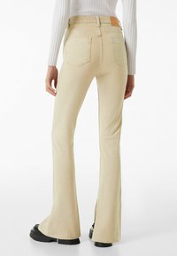 Beiga utsvängda jeans med hög midja, bakfickor och en slitna fåll. Tillverkade av mjukt, stretchigt material; har en brun läderetikett.