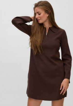 Femme aux longs cheveux portant une robe marron à manches longues avec un col et une fermeture éclair, posant avec une main derrière la tête.