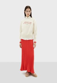 Cremefarbener Pullover mit "LOVE" in roter Schrift, kombiniert mit einem langen, plissierten roten Rock und beigefarbenen, flauschigen Hausschuhen. Lässige Passform, geschichteter Look.