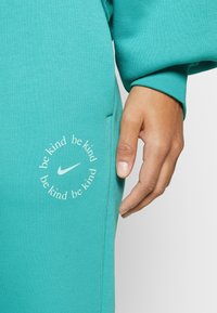 Turquoise sweatshirt with a textured finish. Features a circular white print that says "be kind" around the Nike swoosh logo on the side.