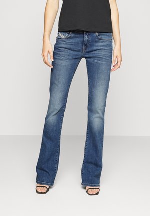 Kobieta ubrana w niebieskie dżinsy bootcut, czarny top i czarne sandały z odkrytymi palcami, stoi na jasnym, jednolitym tle.
