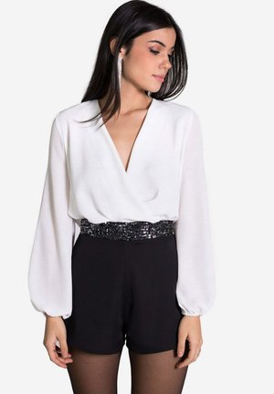 Chemisier blanc à manches longues avec un décolleté en V profond, accompagné de shorts noirs. La taille est soulignée par une ceinture ornée de sequins noirs.