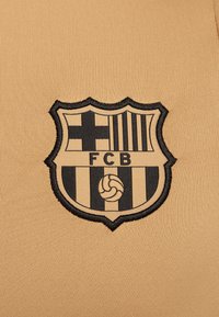 Kangas bež boje sa izvezenim crnim grbom FC Barcelone, sa vertikalnim prugama i centralnim dizajnom fudbalske lopte.