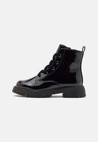 Botín negro de charol con cordones, suela gruesa y lengüeta en un fondo liso gris claro.