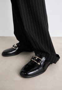 Black leather shoes with a glossy finish, featuring a large gold buckle and strap, paired with pinstriped black trousers.