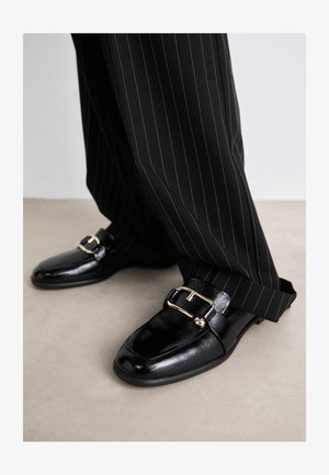 Czarne skórzane buty o połysku, z dużą złotą klamrą i paskiem, zestawione z czarnymi spodniami w paski.