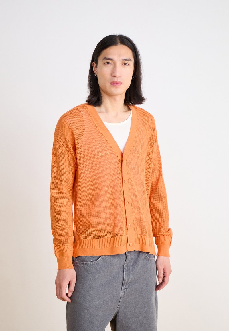 Oranger offener Strickcardigan mit V-Ausschnitt, langen Ärmeln und Knopfverschluss. Über einem weißen Hemd getragen, kombiniert mit grauen Hosen.