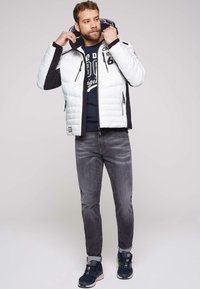 Giacca imbottita bianca e nera con chiusura a zip, tasche laterali e cappuccio, indossata sopra una felpa blu navy e jeans grigi con risvolti.