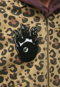 Ζακέτα με φερμουάρ σε μοτίβο λεοπάρ σε ταμπά απόχρωση, με κείμενο μαύρο και άσπρο μπαλώμα φλαμίνγκο στο στήθος, και εσωτερική επένδυση με μπορντό κουκούλα.