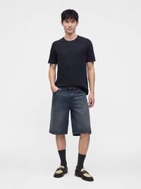 Homme debout, mains partiellement dans les poches, portant un t-shirt noir, un short en jean longueur genou avec une chaîne, des chaussettes noires et des chaussures slip-on noires et beige.