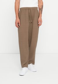 Brown, textured, loose-fitting pants with a drawstring waist. Worn with white sneakers, the fabric appears lightweight and breathable.
