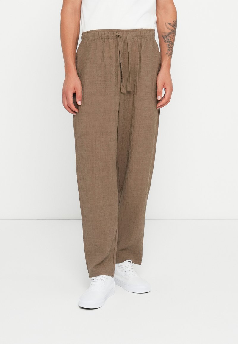 Brown, textured, loose-fitting pants with a drawstring waist. Worn with white sneakers, the fabric appears lightweight and breathable.