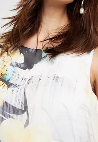 Kwiatowa bluzka w lekkim, zwiewnym materiale. Posiada białą bazę z żółtymi i czarnymi wzorami kwiatowymi, okrągły dekolt oraz luźny krój.