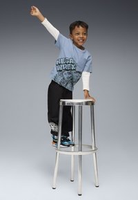 Jaunas berniukas šypsosi stovėdamas ant aukšto metalinio tabureto, viena ranka pakelta, o kita laiko taburetę.